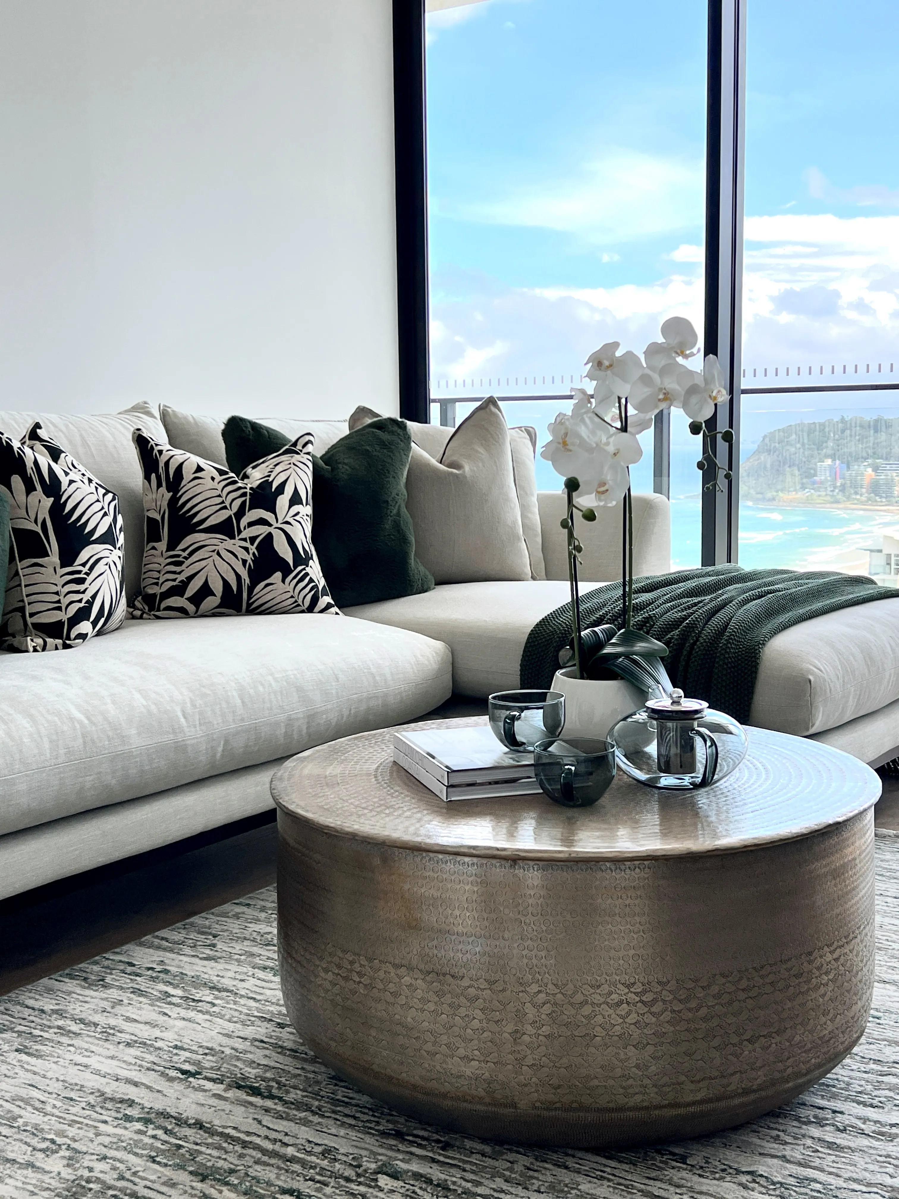Media room with cream sofa, deep green and botanical cushions, hammered brass coffee table and ocean views